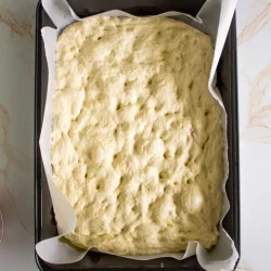 Un molde para hornear rectangular forrado con papel pergamino contiene masa de focaccia cruda.