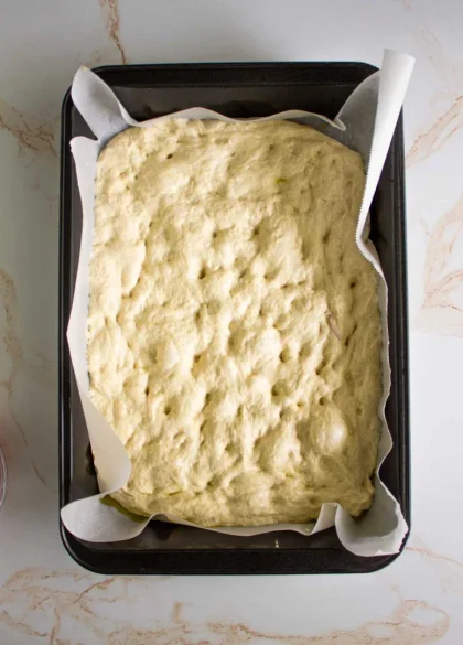 Un molde para hornear rectangular forrado con papel pergamino contiene masa de focaccia cruda.