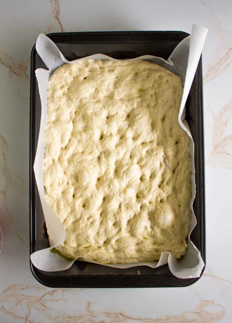Un molde para hornear rectangular forrado con papel pergamino contiene masa de focaccia cruda.