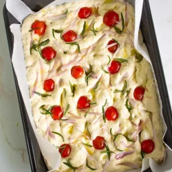 Una bandeja de masa de focaccia sin hornear cubierta con tomates cherry cortados por la mitad, ramitas de romero y chalotas en juliana. La masa se coloca sobre papel de pergamino en una bandeja para hornear rectangular.
