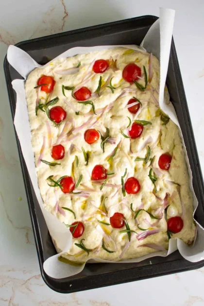 Una bandeja de masa de focaccia sin hornear cubierta con tomates cherry cortados por la mitad, ramitas de romero y chalotas en juliana. La masa se coloca sobre papel de pergamino en una bandeja para hornear rectangular.