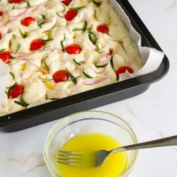 Una bandeja de horno con masa de focaccia cruda cubierta con tomates cherry, romero y cebollas rojas. Al lado se coloca un pequeño recipiente de vidrio con aceite de oliva emulsionado.