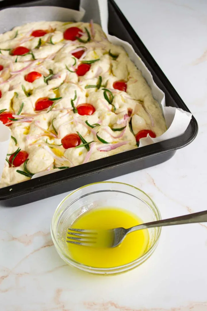 Una bandeja de horno con masa de focaccia cruda cubierta con tomates cherry, romero y cebollas rojas. Al lado se coloca un pequeño recipiente de vidrio con aceite de oliva emulsionado.