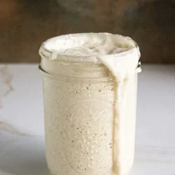 A glass jar filled with bubbling sourdough starter overflows slightly onto a marble surface.