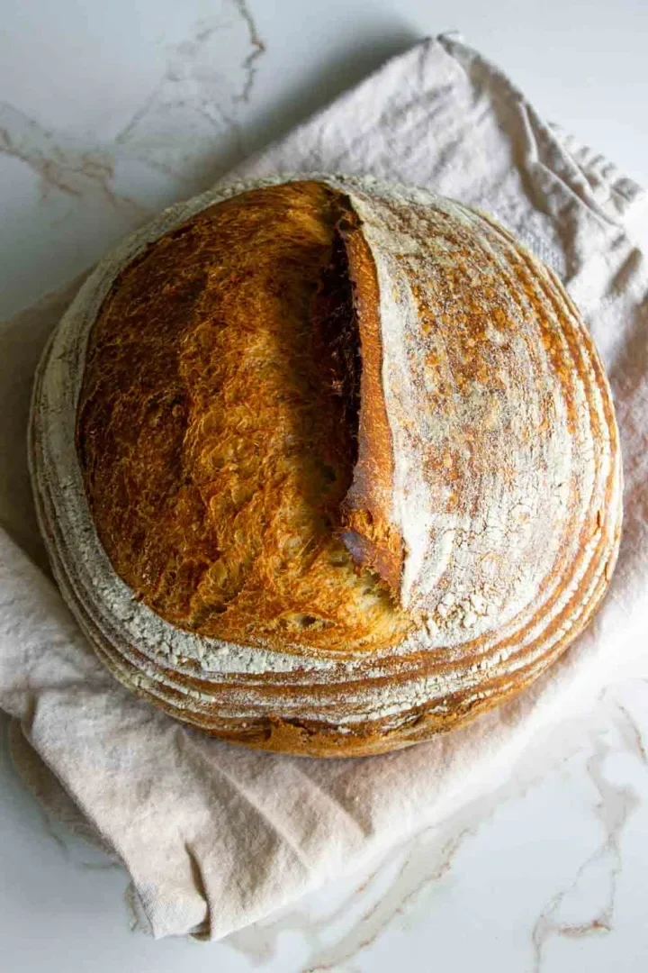 Round loaf of crusty sourdough bread with a golden-brown crust, resting on a light cloth.