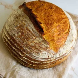 A round, rustic loaf of sourdough bread with a golden crust and flour dusting rests on a light cloth.