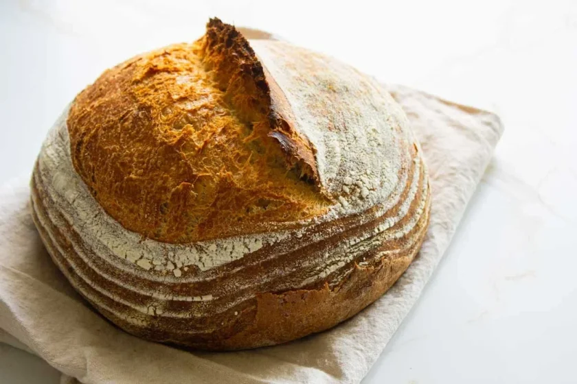 Round loaf of crusty sourdough bread on a beige cloth, with a split top and a flour-dusted surface.