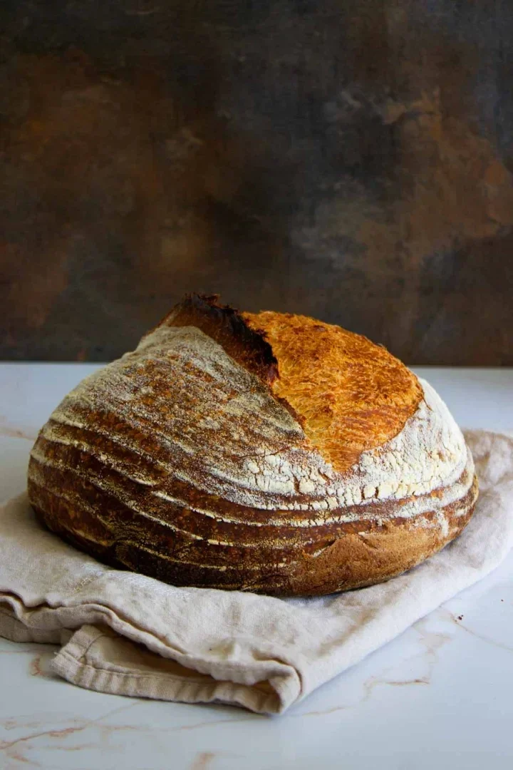 Round loaf of rustic sourdough bread with a cracked, golden crust, resting on a beige cloth against a dark, textured background.