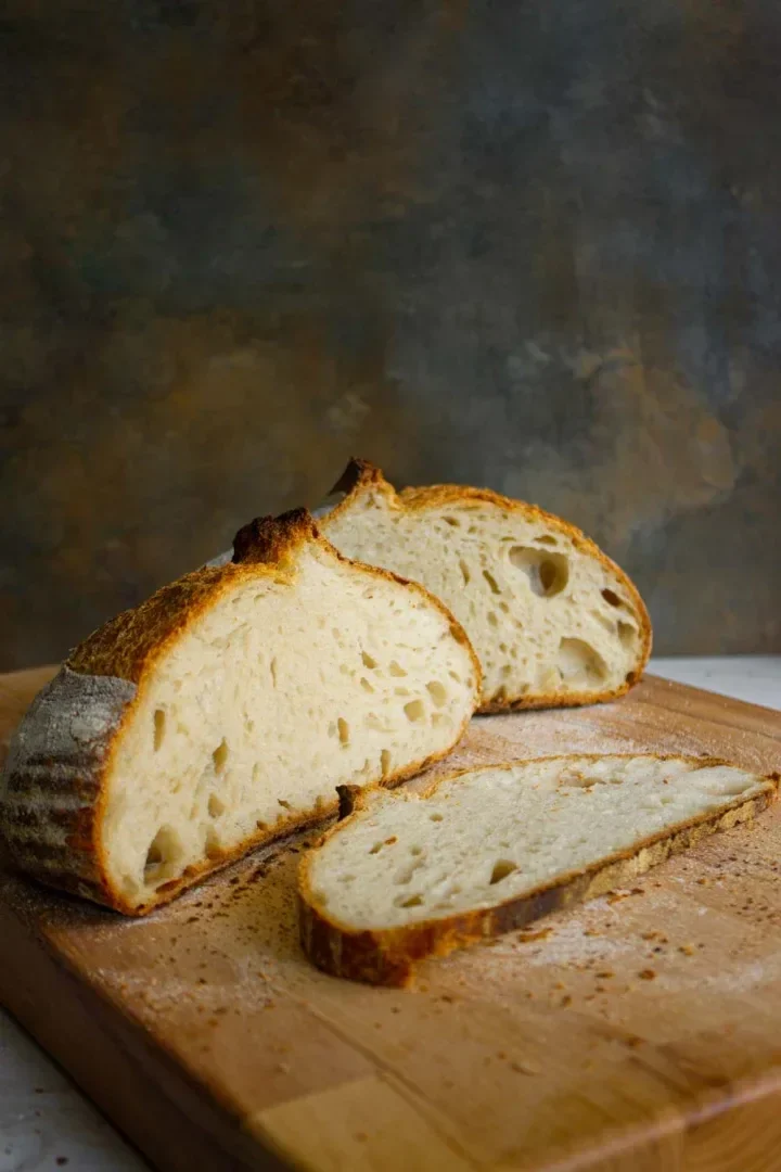 A sliced loaf of rustic sourdough bread on a wooden cutting board, with one piece laid flat.