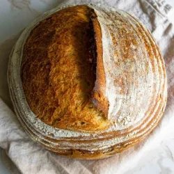 Una hogaza redonda de pan crujiente de masa madre, horneado y con la parte superior abierta, reposa sobre un paño colocado sobre una superficie de mármol.