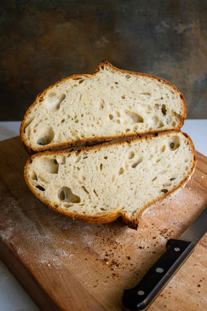 Pan de masa madre cortado en rodajas con un exterior crujiente y un interior aireado sobre una tabla de cortar de madera, acompañado de un cuchillo.