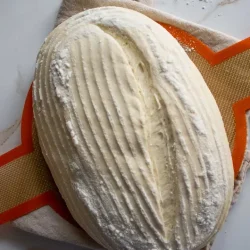 Una hogaza de masa de pan sin hornear sobre una estera de silicona naranja texturizada, sobre un paño beige, y sobre una superficie de mármol. La masa tiene un patrón rayado y enharinado.