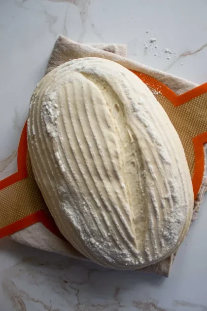 Una hogaza de masa de pan sin hornear sobre una estera de silicona naranja texturizada, sobre un paño beige, y sobre una superficie de mármol. La masa tiene un patrón rayado y enharinado.