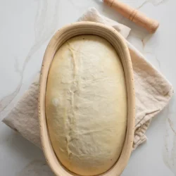 Oval-shaped dough resting in a proofing basket with a dough scraper on a cloth nearby, placed on a marble surface.