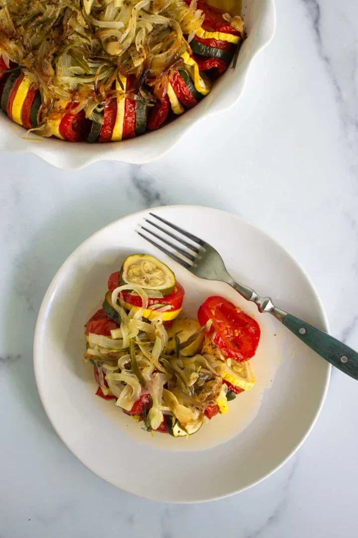 A serving of roasted vegetables, including zucchini, tomato, and yellow squash, topped with caramelized onions on a white plate with a fork; a baking dish with more vegetables is visible above.
