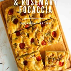 A wooden cutting board holds sliced cherry tomato and rosemary focaccia bread. TText on the image reads "Cherry Tomato & Rosemary Focaccia, No Knead Recipe.