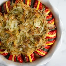 A round baking dish filled with neatly arranged slices of zucchini and tomato, topped with sautéed onions and herbs, set on a white marble surface.