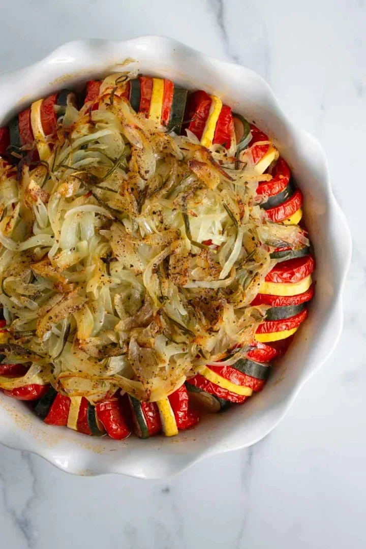 A round baking dish filled with neatly arranged slices of zucchini and tomato, topped with sautéed onions and herbs, set on a white marble surface.