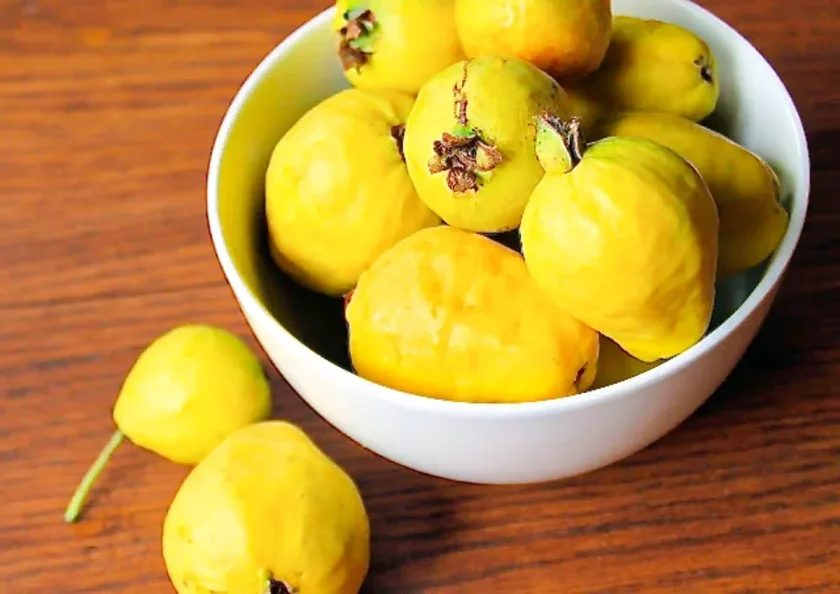 A white bowl filled with ripe yellow guavas sits on a wooden surface, with two guavas beside it.