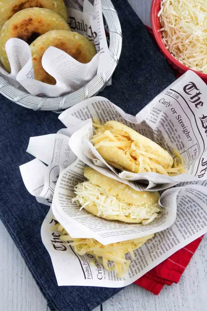 Tres arepas rellenas de queso están envueltas en papel estilo periódico, con más arepas y un tazón de queso rallado en el fondo.