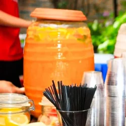 Large jar of rum guarapita with lemon slices, plastic cups, straws, and napkins on an outdoor table.