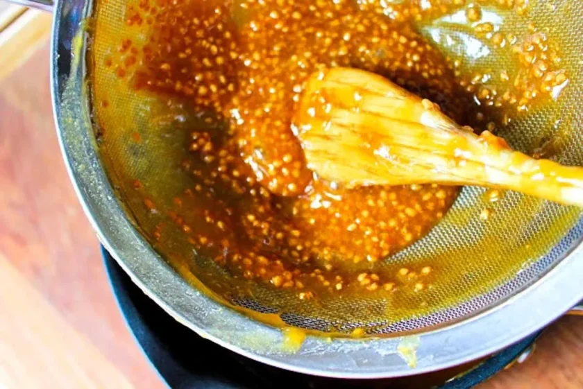 A wooden spoon stirs a guava jam mixture in a fine mesh strainer over a bowl.