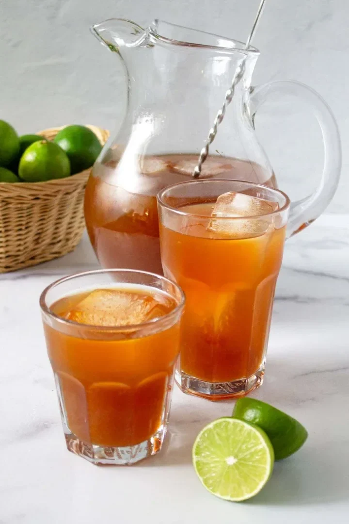 Una jarra de cristal y dos vasos de guarapo de papelón con limón con cubitos de hielo se encuentran sobre una superficie blanca. Al fondo, una lima cortada y una cesta de limas enteras.