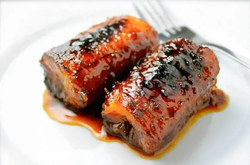 Dos piezas de panceta de cerdo braseada glaseada con superficie caramelizada servidas en un plato blanco con un tenedor en el fondo.