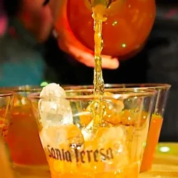 Una mano con una botella de guarapita de ron y parchita, la sirve en un vaso desechable con hielo. En el vaso se lee Santa Teresa.