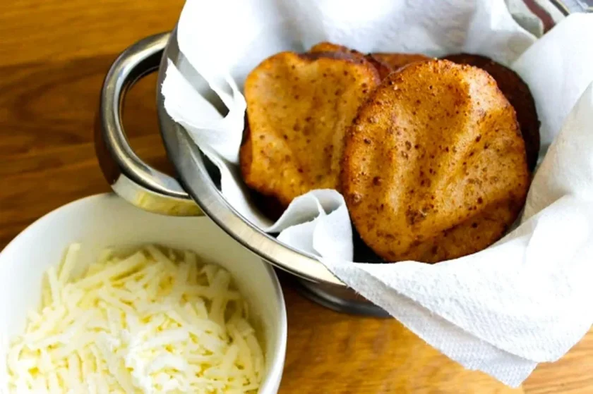 Un cuenco de metal con arepas de papelón y anís fritas sobre papel absorbente junto a un cuenco de queso blanco rallado.