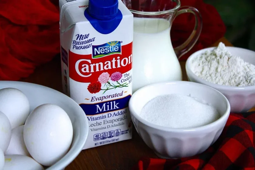 Eggs, evaporated milk, sugar, flour, and a milk pitcher over a plaid black and red tablecloth.