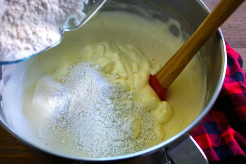 Harina añadida a un bol de masa de torta tres leches con una espátula sobre un paño rojo y negro.