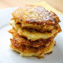 Una pila de tortitas de patata doradas y crujientes en un plato blanco, que recuerda al pan francés, ligeramente espolvoreadas con azúcar en polvo.