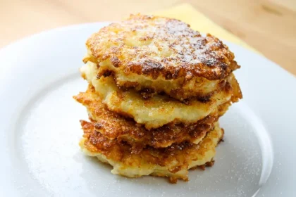 Una pila de tortitas de patata doradas y crujientes en un plato blanco, que recuerda al pan francés, ligeramente espolvoreadas con azúcar en polvo.