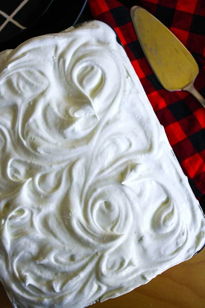 Una torta tres leches cubierta de merengue en espiral junto a un mantel de cuadros rojos y negros y un servidor de tartas.