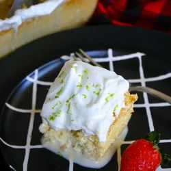 Un trozo de tres leches con cobertura de merengue con ralladura de lima, en un plato negro, servido con una fresa fresca.