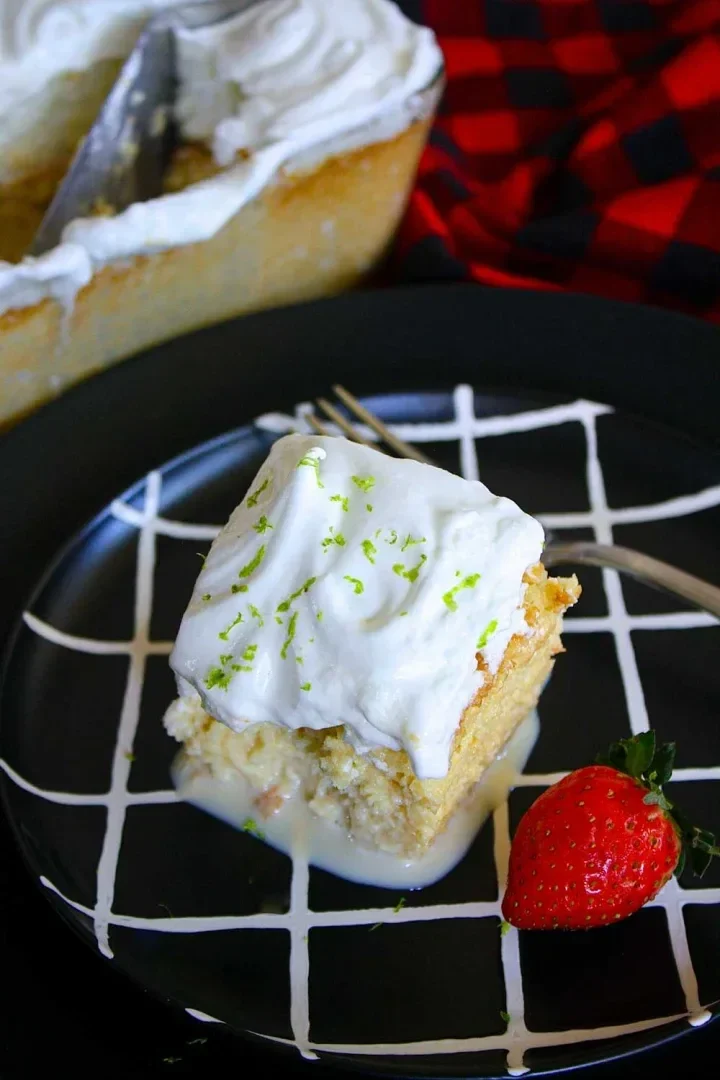 Un trozo de tres leches con cobertura de merengue con ralladura de lima, en un plato negro, servido con una fresa fresca.
