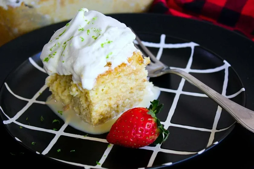 Un trozo de torta tres leches con merengue y ralladura de lima, servido con una fresa en un plato negro cuadriculado.