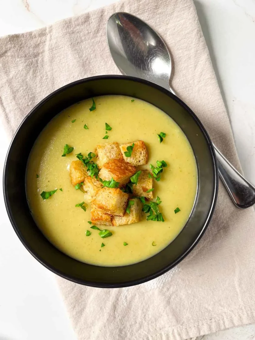 Un cuenco de sopa cremosa de ajo porro y papa cubierto con crutones y un poco de perejil picado, junto a una cuchara sobre una servilleta beige.