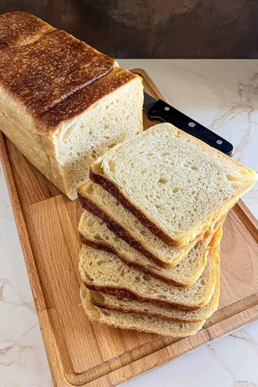 Una hogaza de pan de molde de masa madre descansa sobre una tabla de cortar de madera, con un cuchillo al lado, lista para ser degustada.