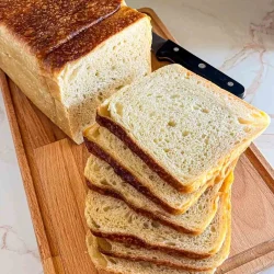 Una hogaza de masa madre pan de sándwich con varias rebanadas cortadas, colocada sobre una tabla de cortar de madera con un cuchillo.