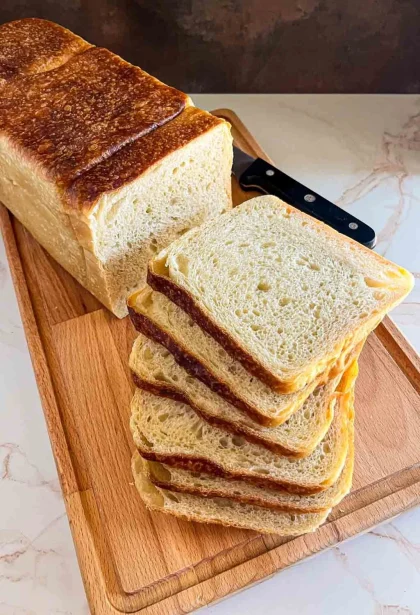 Una hogaza de masa madre pan de sándwich con varias rebanadas cortadas, colocada sobre una tabla de cortar de madera con un cuchillo.