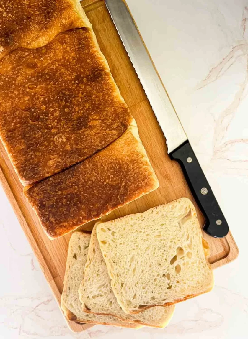 Una barra de pan sobre una tabla de cortar de madera con un cuchillo de pan y tres rebanadas de pan.