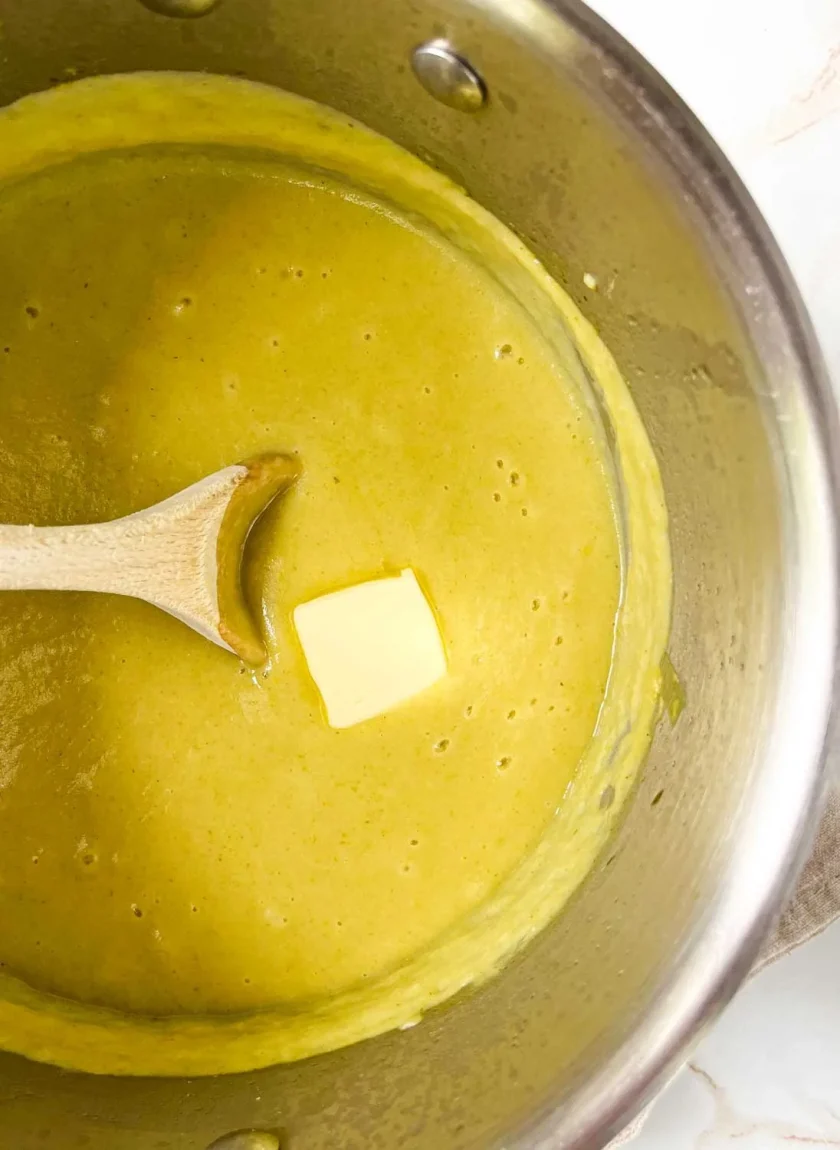 Una cuchara de madera en una olla de cremosa sopa amarilla con una palmadita de mantequilla derritiéndose por encima. La sopa de es poro con papa.