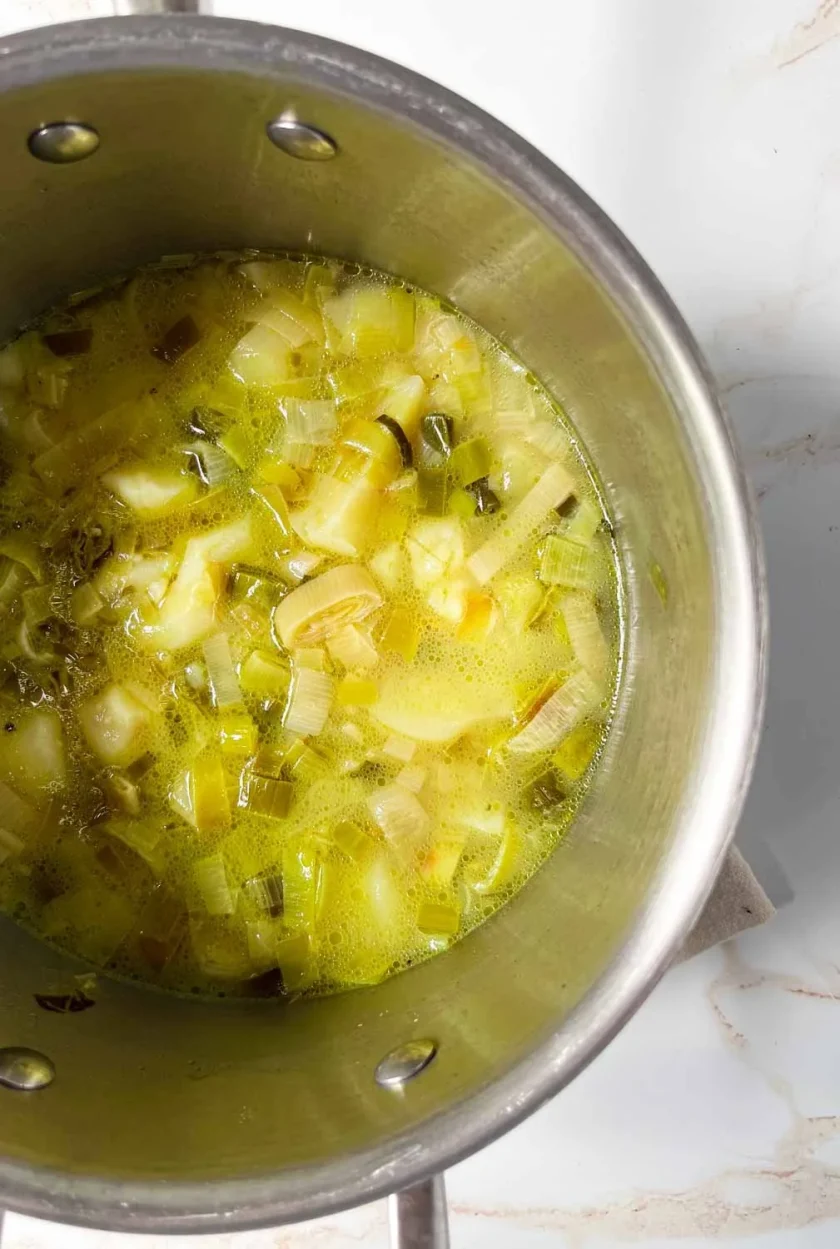 Una olla de sopa de puerros picados, patatas y caldo sobre una superficie blanca.
