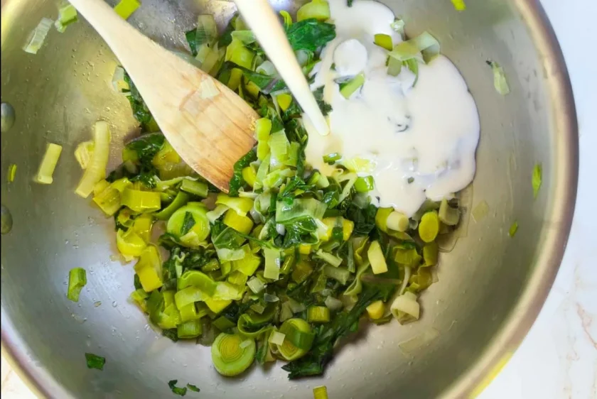 Puerros y verduras picados en una sartén con una cuchara de madera, se mezclan con un chorrito de crema de leche.