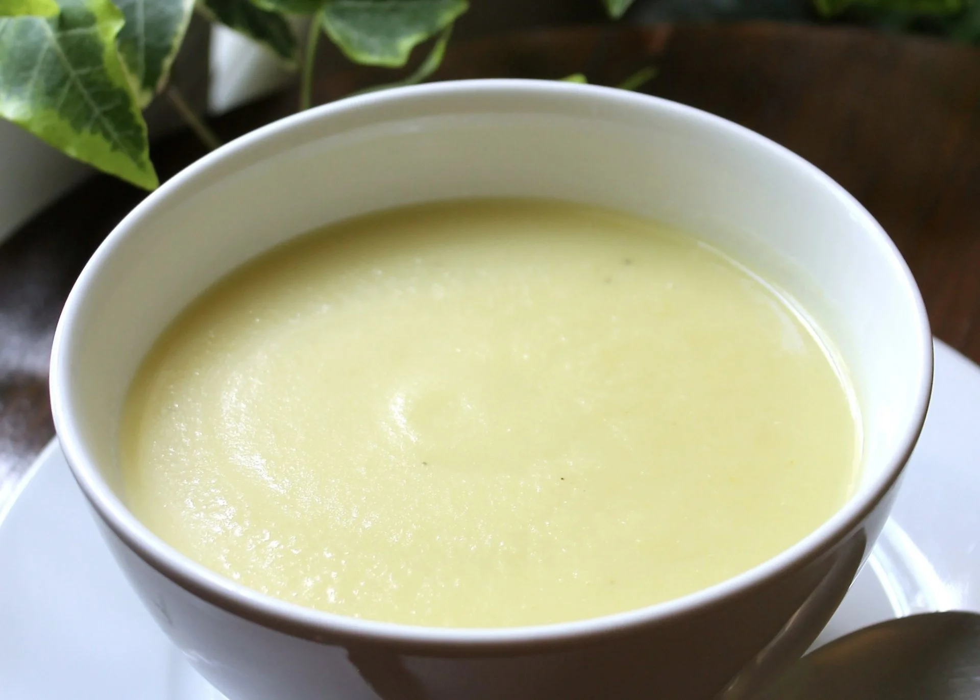 Un cuenco blanco lleno de cremosa y suave sopa amarilla de crema de ajo porro, colocado sobre un platillo blanco.