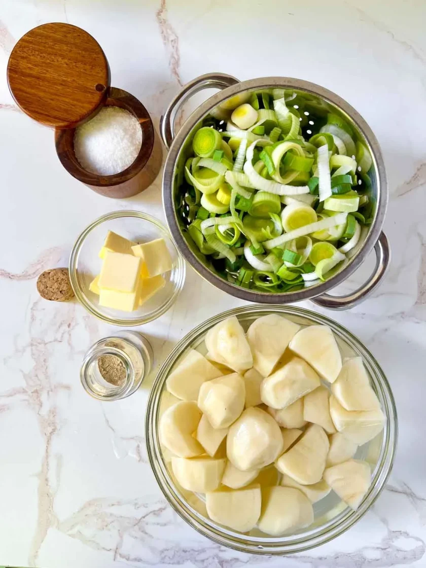 Cuencos con patatas troceadas, puerros en rodajas, mantequilla, sal, pimienta sobre una encimera de mármol.
