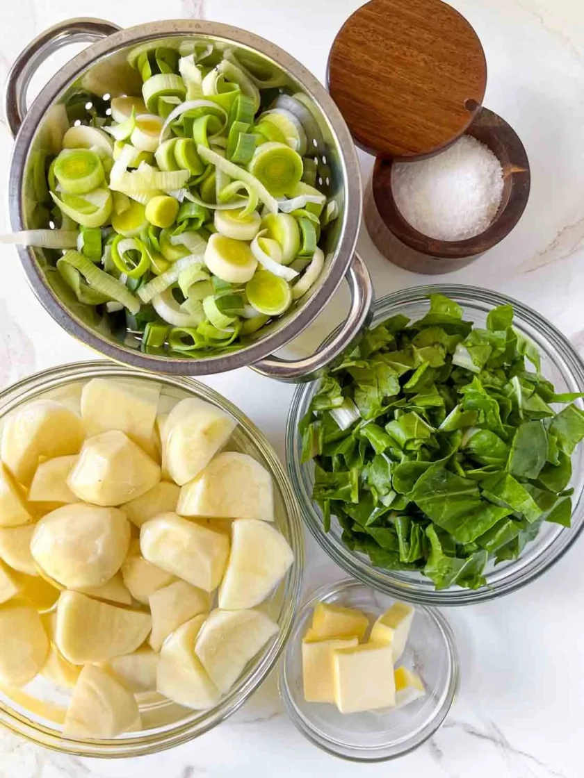 Puerros picados, acelgas y papas en cuencos con mantequilla y sal sobre una superficie de mármol blanco.