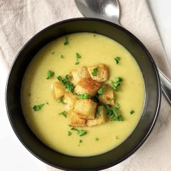 Un cuenco de sopa cremosa de ajo porro con papa, cubierto con picatostes y perejil, junto a una cuchara sobre una servilleta beige.